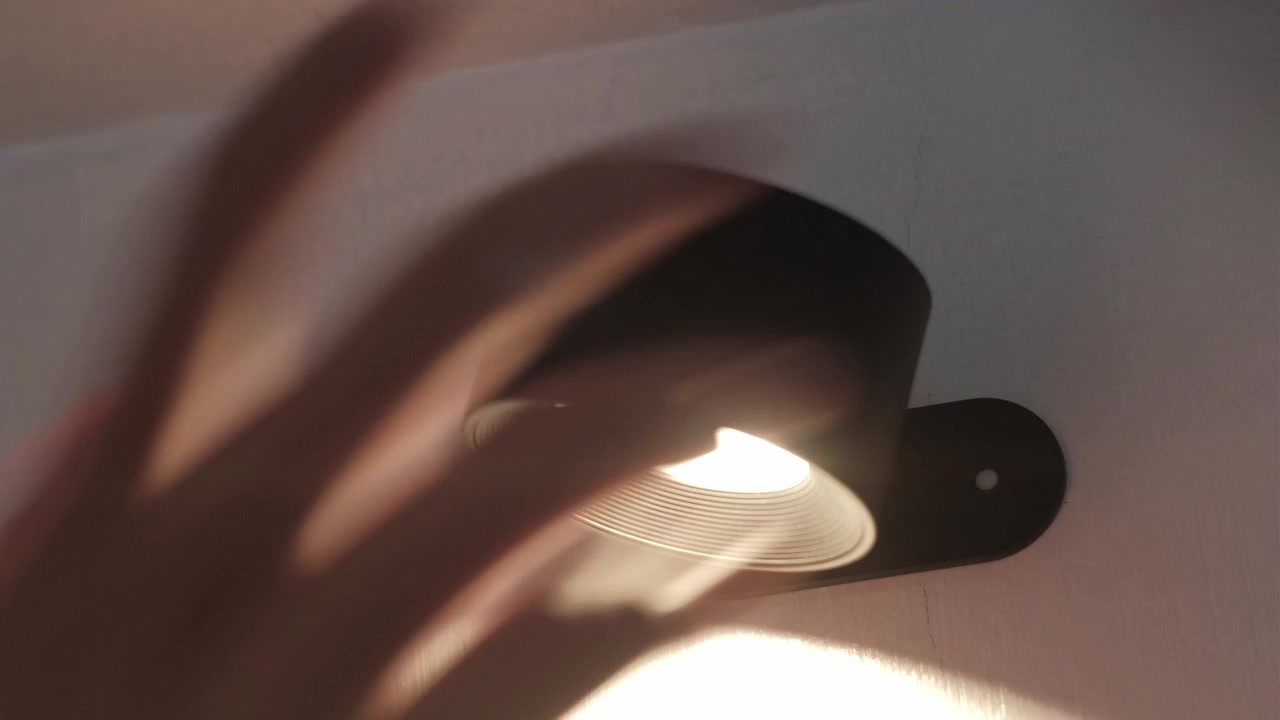 Plano estático aproximado de uma mão fixando e ligando uma luz LED de parede em ambiente interno, revelando uma iluminação quente numa parede branca.