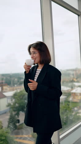 Positive pensive lady holding a cup looks at window. Happy business person having break from work. Vertical video.