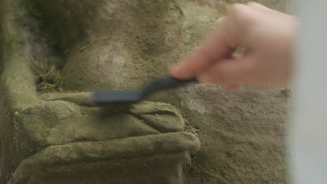 hand using tool to remove vegetation from old stone sculpture