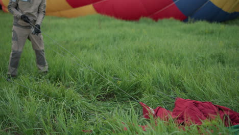 crew member dragging colorful nylon balloon envelope with bright red yellow panels across tall grass field during preparation layout alongside teammate wearing gloves under pastel sky