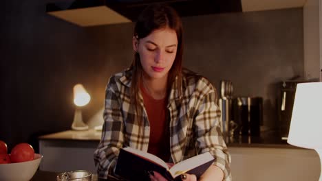 happy brunette girl in a plaid shirt and an orange t-shirt stands in the kitchen and reads a book during her evening relaxation in a modern apartment