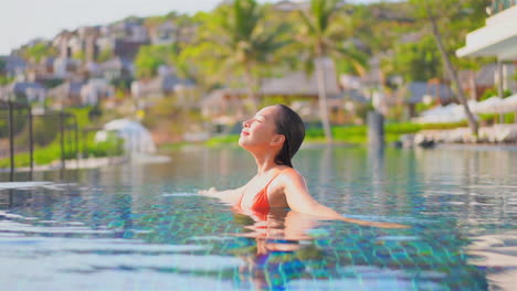 빨간 비키니를 입은 아시아 여성이 그리스의 한 개인 빌라에 있는 수영장 안의 양손에 양손을 펴고 돌아서며 낮에는 눈을 감고 슬로우 모션을 하고 있습니다.