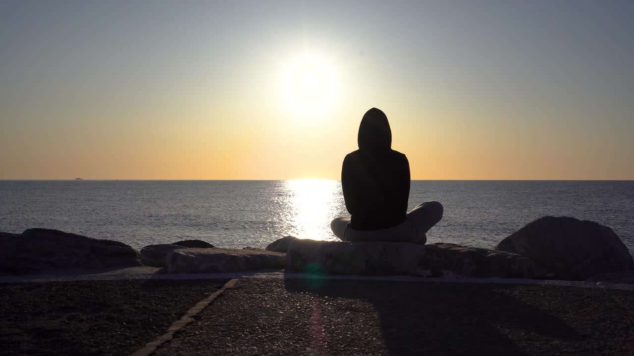 silueta de una chica con capucha disfrutando del amanecer temprano en la mañana en el muelle de puerto banús con vistas al mar mediterráneo, pose de yoga pose de ángulo enlazado, filmada en 4k en gimbal