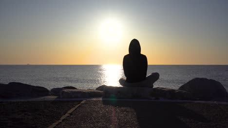 Silueta-De-Una-Chica-Con-Capucha-Disfrutando-Del-Amanecer-Temprano-En-La-Mañana-En-El-Muelle-De-Puerto-Banús-Con-Vistas-Al-Mar-Mediterráneo,-Pose-De-Yoga-Pose-De-ángulo-Enlazado,-Filmada-En-4k-En-Gimbal