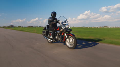 Travel-Dream---Biker-Rides-Along-A-Country-Road-On-A-Background-Of-Blue-Sky-And-Green-Fields