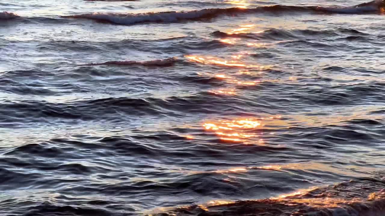 A close-up shot of the ocean surface at dusk. The dark water is covered in gentle ripples and small waves, with the warm, golden light of the setting sun creating brilliant sparkling reflections