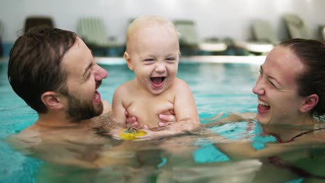 Junge-Mutter-Und-Vater-Halten-Ihr-Süßes-Kind-Im-Schwimmbad-Und-Küssen-Sich-Gegenseitig-Auf-Die-Lippen.-Glückliches-Kind-Lächelt