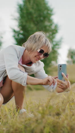 mujer de mediana edad con gafas de sol en cuclillas mientras toma una foto con una cálida sonrisa en tierras de cultivo abiertas con árboles en el fondo, la luz natural destaca su expresión alegre