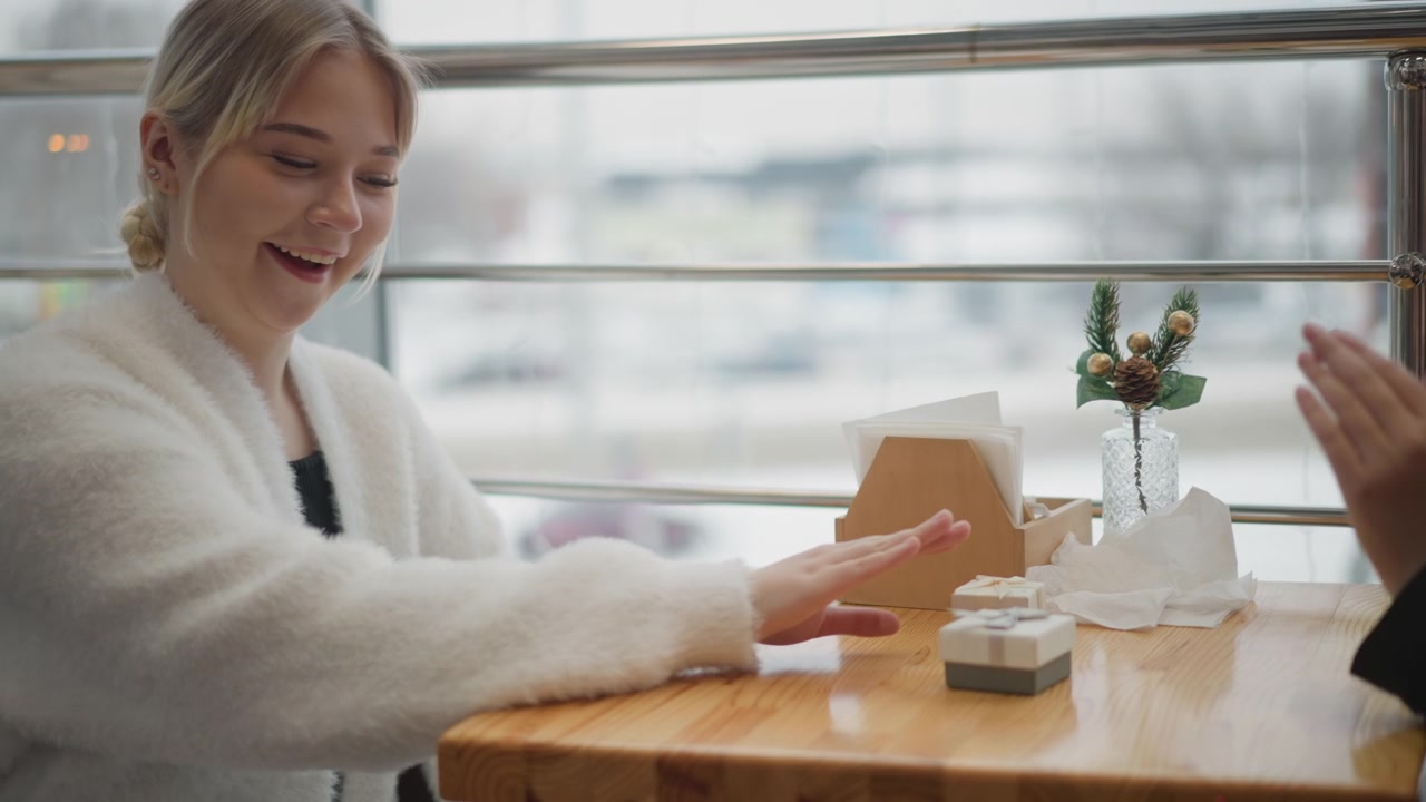Smilende kvinde i hvid sweater sidder ved et træcafébord og rækker frem og giver en to-go kop til sin ledsager, hvilket viser afslappet social interaktion, blid håndbevægelse og en varm indendørs café