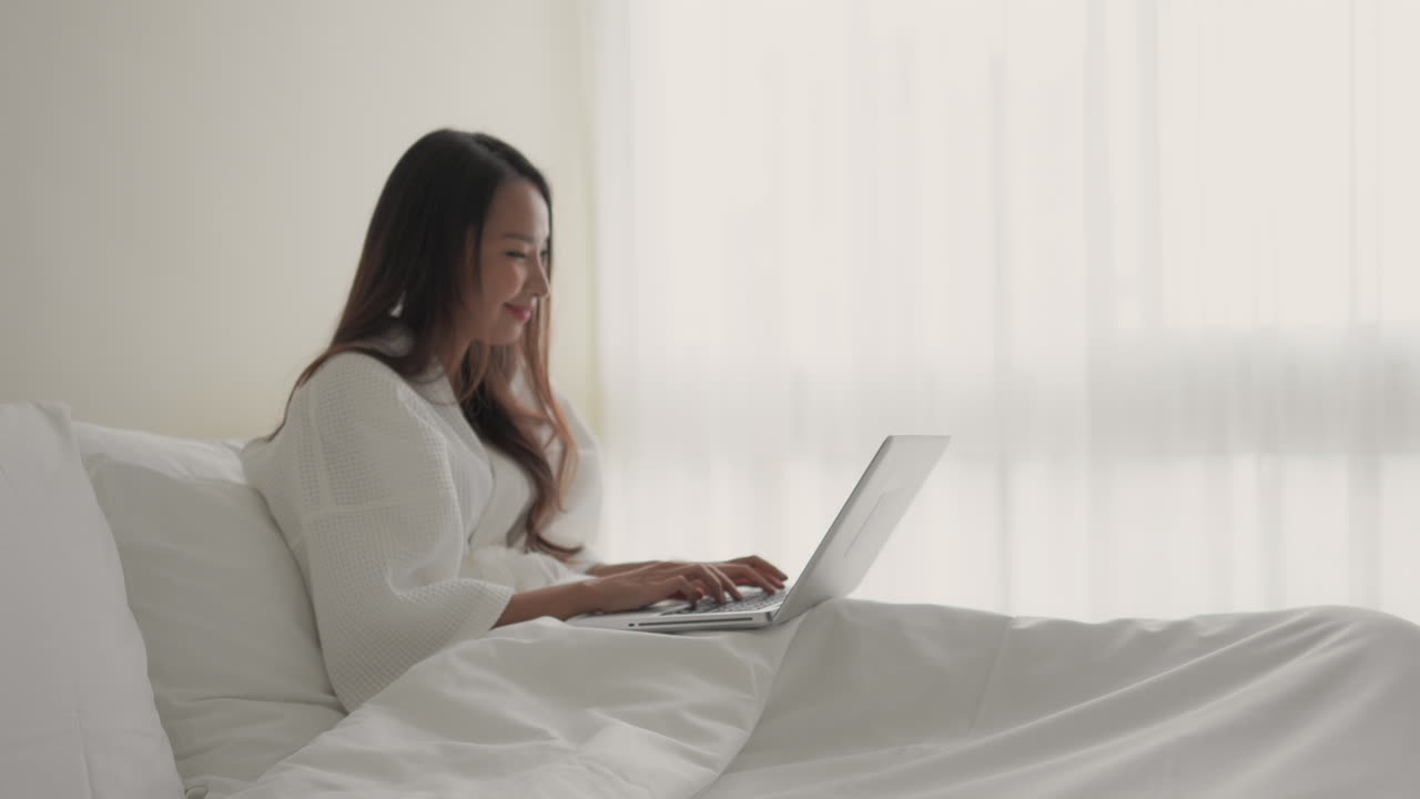 침대에 누워 있는 예쁜 아시아 여성이 노트북에 타이핑을 하는 동안 베개에 기대어 있다