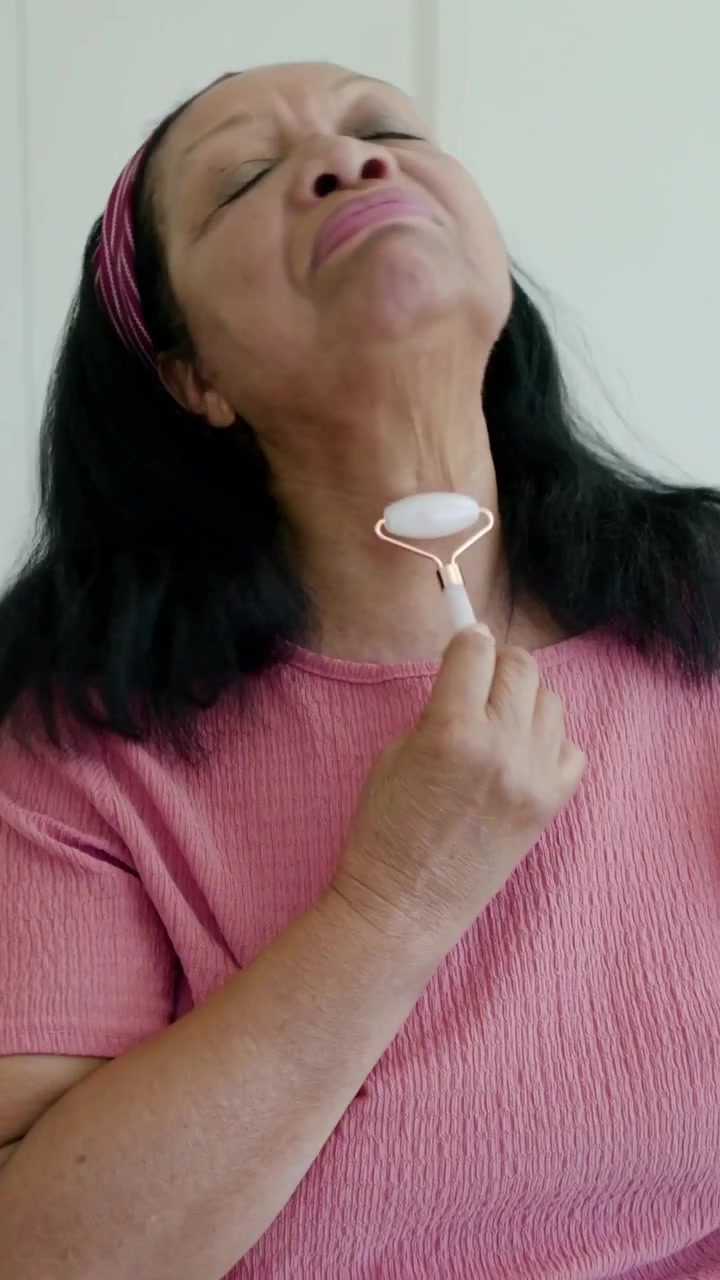 Video verticale: Donna che solleva un rullo rosa e poi lo passa sulla mascella e sul collo per la cura della pelle, vicino all'armadio.