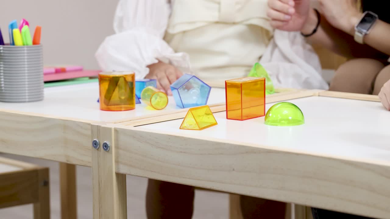 Two people manipulate translucent geometric solids on a wooden table in bright, indoor classroom lighting