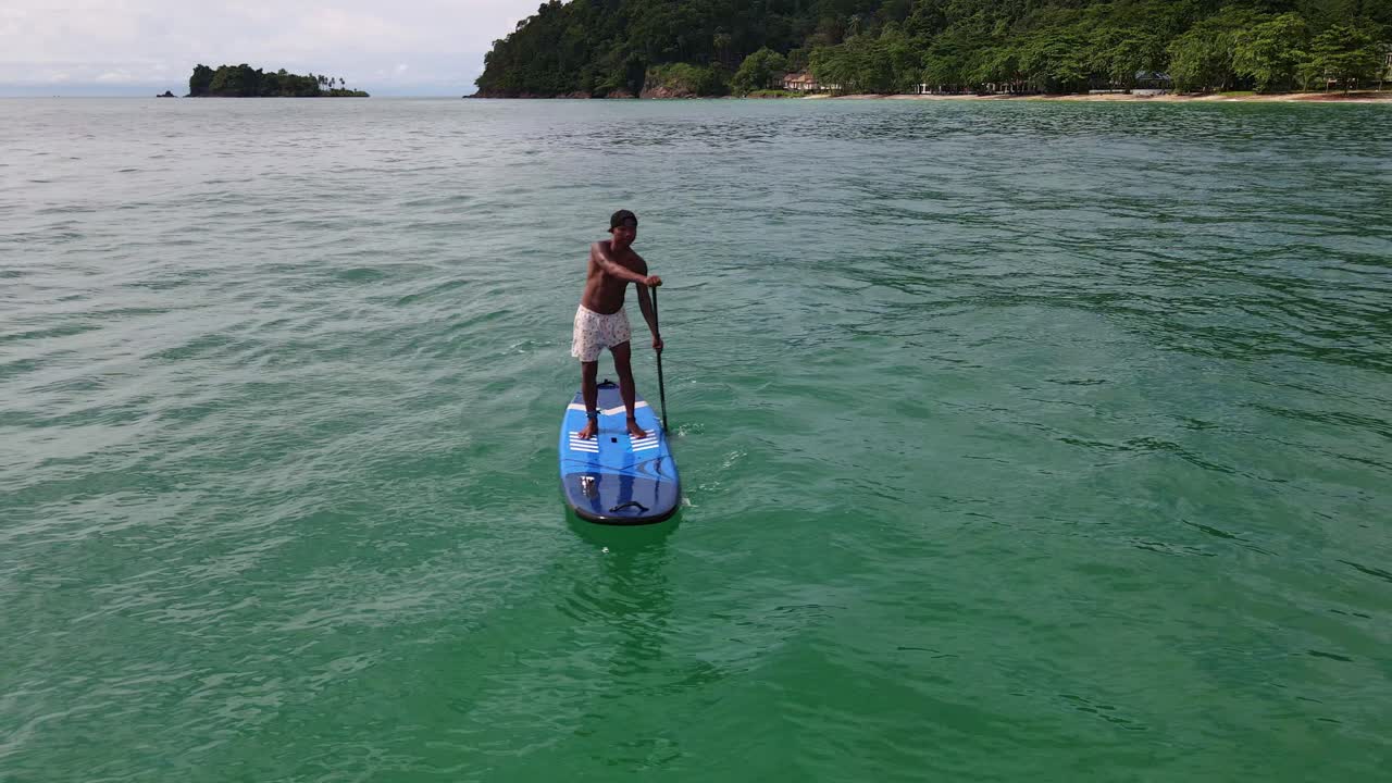 Ripresa aerea con drone da angolazione bassa di un uomo asiatico che si esercita su una tavola da SUP in acque tropicali turchesi e cristalline, con spiaggia, isole e costa in Thailandia.