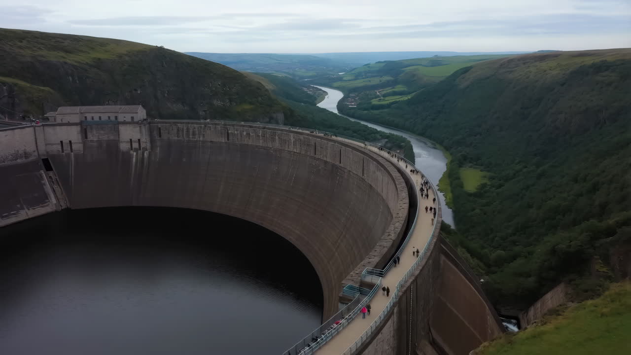 Panoramic view of a large concrete dam with people walking on its crest, overlooking a river valley