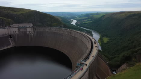 panoramic view of a large concrete dam with people walking on its crest, overlooking a river valley