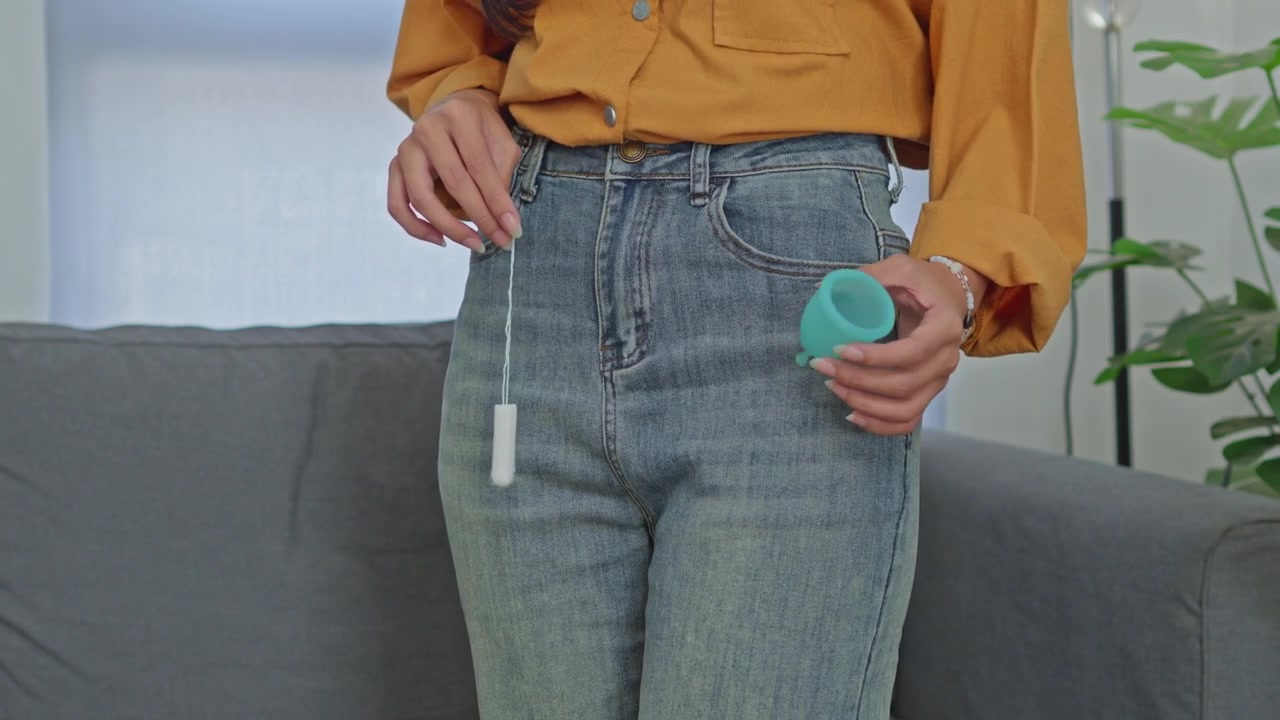 Midsection of woman holding cotton tampon and green menstrual cup in front of her jeans at home