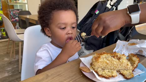 Adorable-and-exotic-two-year-old-child-eating-a-cake-in-a-cafeteria-with-mum,-wearing-a-white-sleeveless-shirt