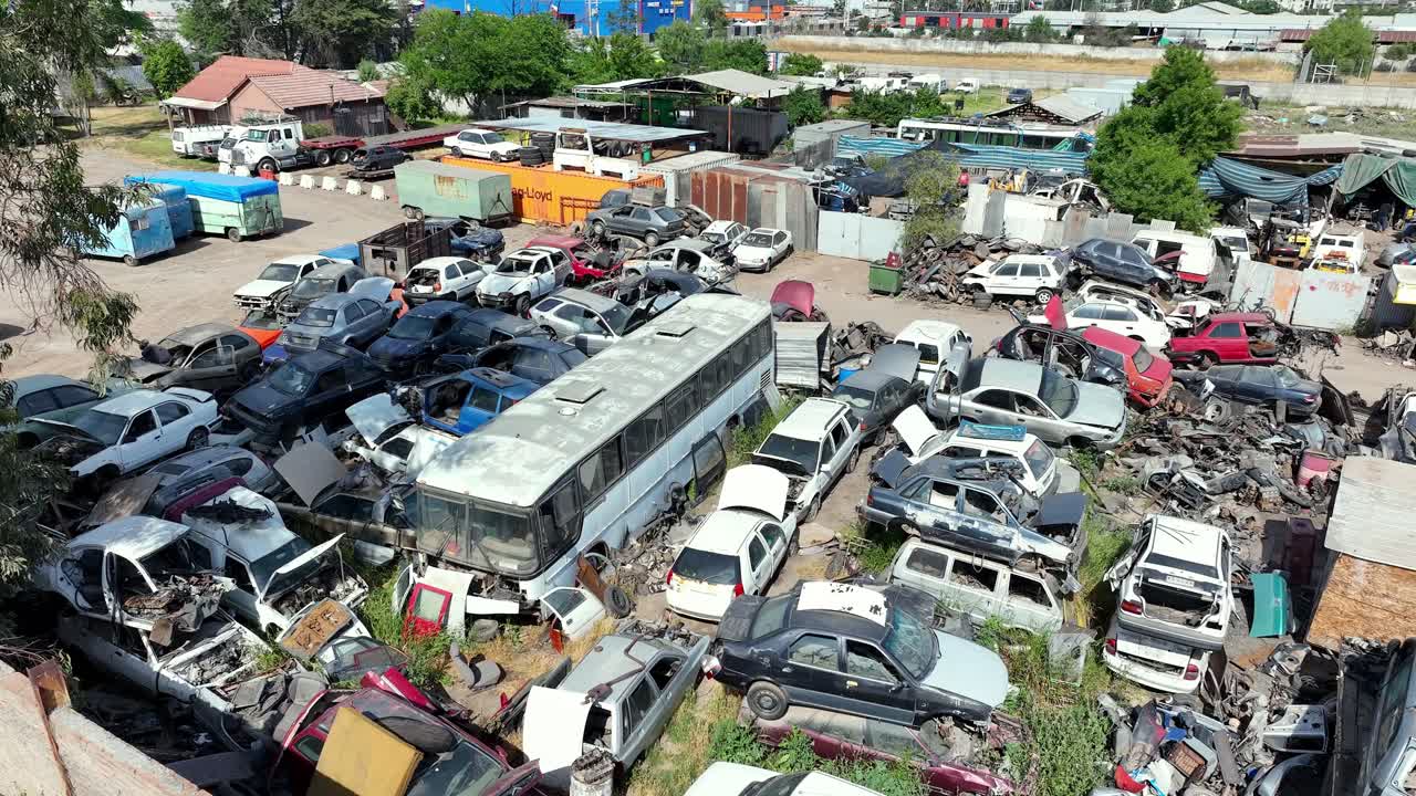 Orbiting drone aerial of chaotic junkyard with scrapped cars, bus, trucks, trailers, and auto parts in industrial salvage yard