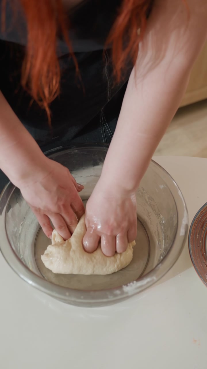 Close top view of both hands kneading soft dough inside clear glass bowl on light kitchen countertop, showing pressing, folding, stretching, and shaping during homemade baking preparation