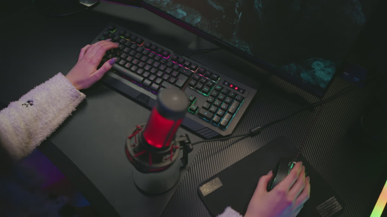top down view of woman wearing fluffy sweater playing video game using colorful keyboard and mouse with red microphone placed on dark desk in stylish gaming studio with glowing monitor and LED lights