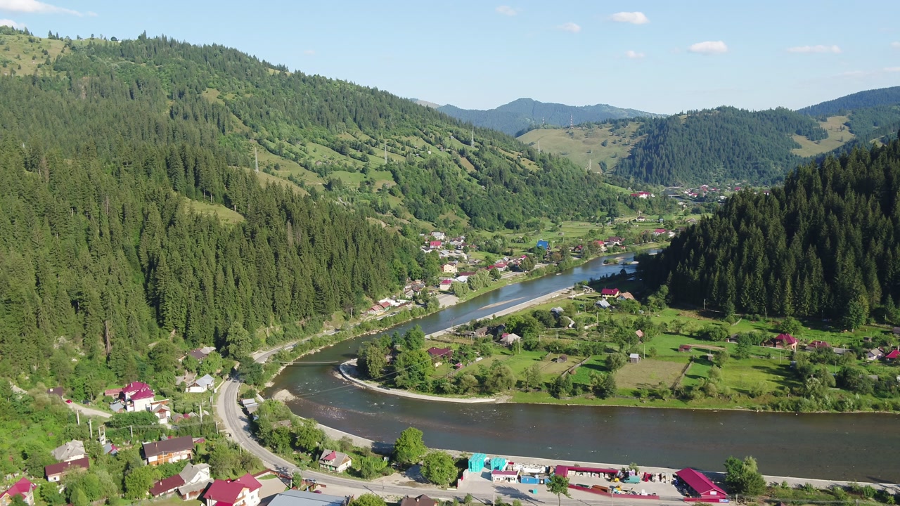 A high-angle aerial shot slowly orbits over a rural village in a Romanian mountain valley. Houses are scattered along a wide river bend, surrounded by lush green summer forests. Brosteni, Suceava.