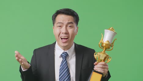 close up of wondering asian business man in a suit and tie with a gold medal looking at trophy doubtfully on green screen background in the studio