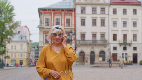 Senior-old-tourist-woman-in-stylish-clothes-talking-on-mobile-phone-while-walking-on-city-street