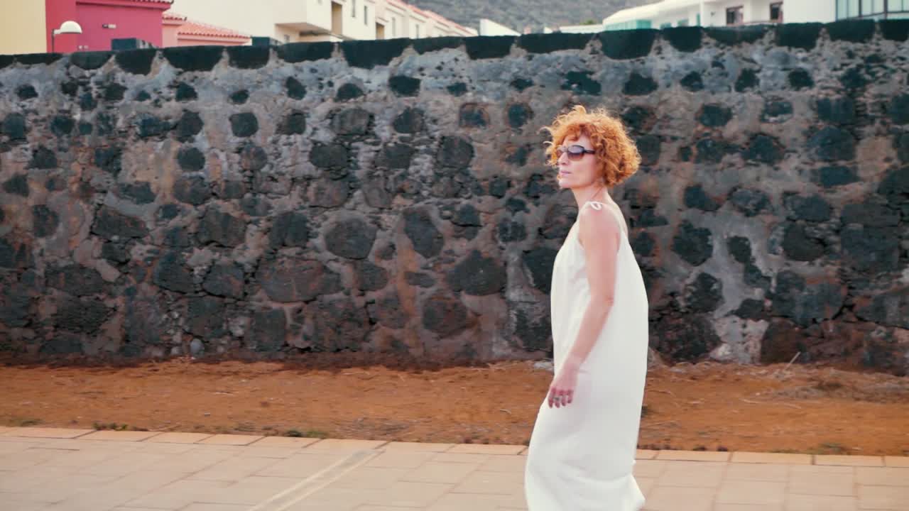 바람 부는 날에 검은 벽을 따라 걷는 흰색 옷을 입은 빨간 머리 여자