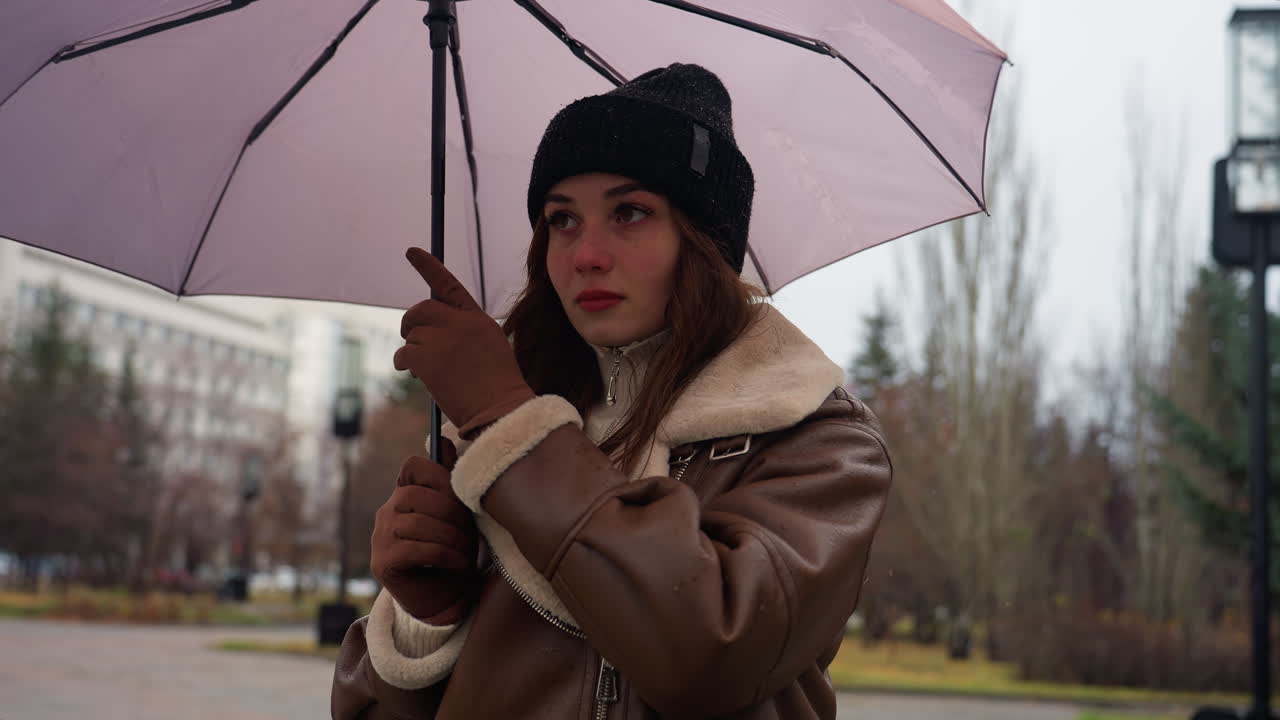 Thoughtful lady holding umbrella in cold weather, wearing black knit cap and brown shearling jacket, standing outdoors surrounded by pine trees on overcast day with soft light snowfall