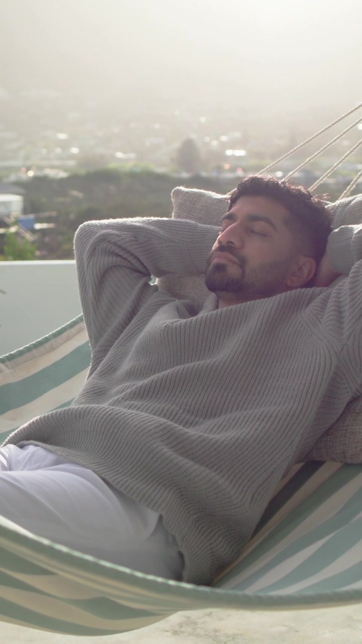 Video verticale: Uomo sdraiato con maglione grigio che riposa su un balcone, la telecamera panoramica rivela un'amaca a strisce.