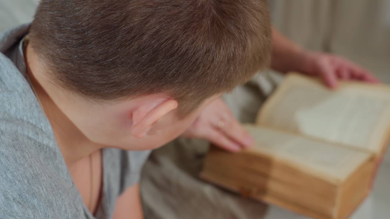 side view of boy in grey shirt intently looking into old worn book while partial view of woman flipping fragile page, soft indoor light highlighting texture of aged paper and thoughtful atmosphere