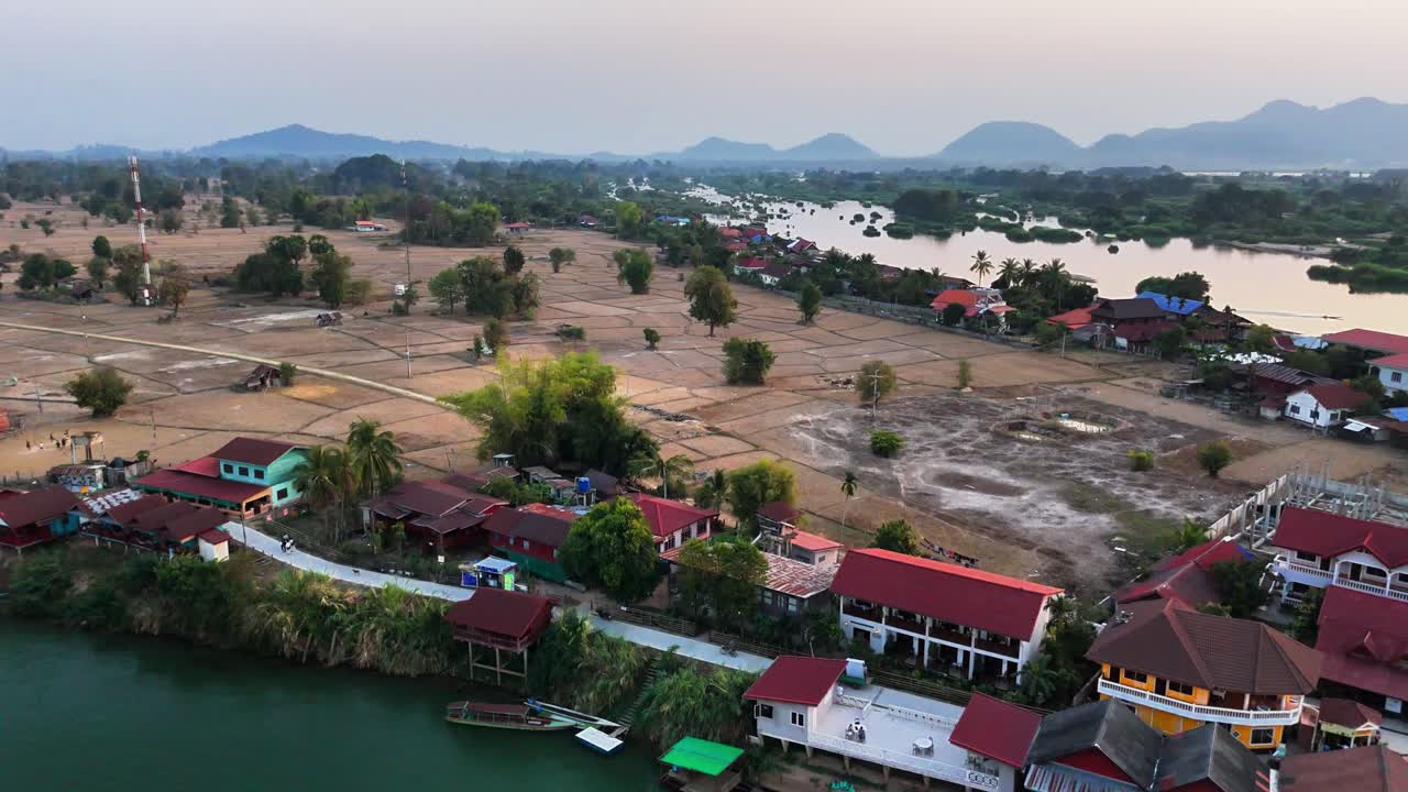 Drone captures rice paddies and village homes in Don Det, Laos, bordered by the Mekong River and backed by the distant Annamite Mountains, as local farming and land preparation unfold at dusk