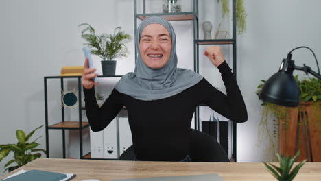 woman in hijab smiling and looking at her phone while sitting at her desk in the office