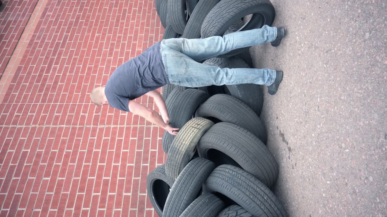 Caucasian male stacking old used tires in front of a brick wall to be recycled, vertical