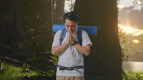 excursionista masculino asiático con mochila de montañismo rezar por algo mientras explora la naturaleza del bosque