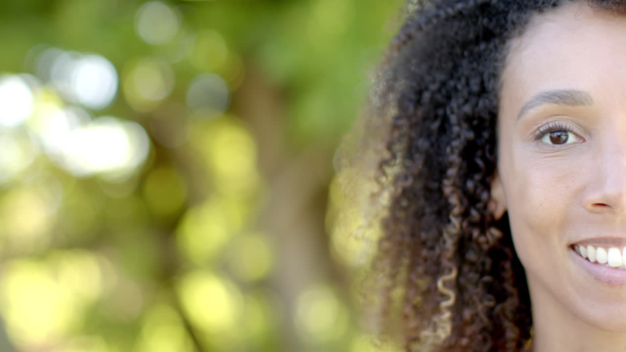 Meio retrato de mulher birracial com cabelo cacheado sorrindo em jardim ensolarado, em câmera lenta, com espaço para texto.