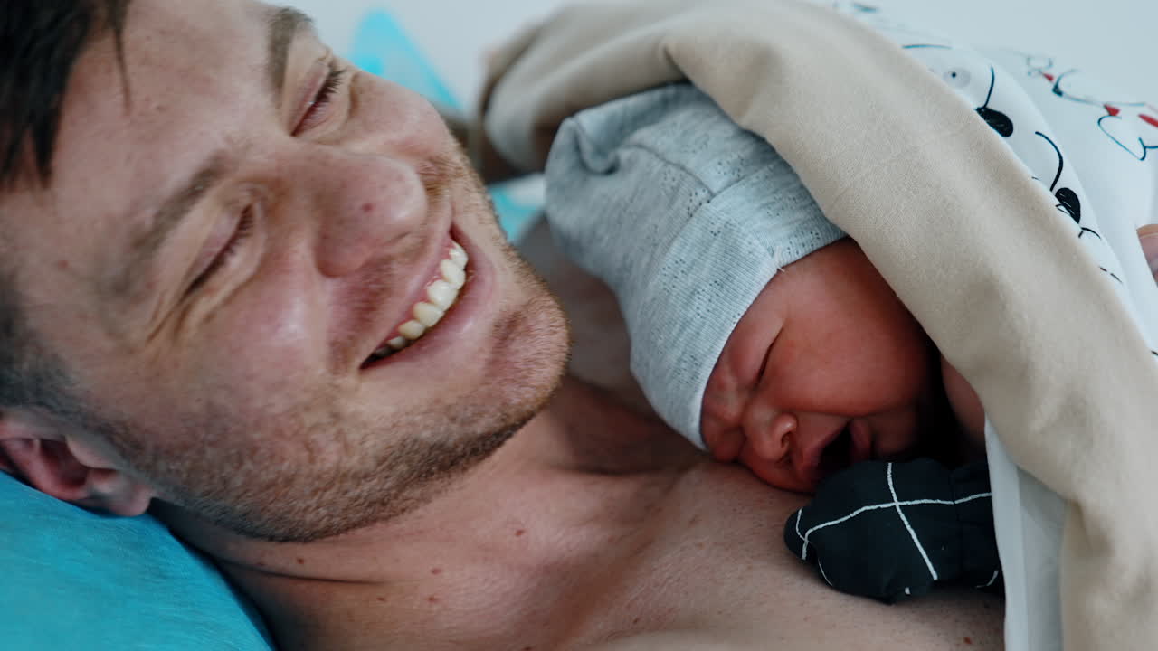 Crying newborn baby lying on the dad's chest after birth. Happy father smiles to camera. Close up.