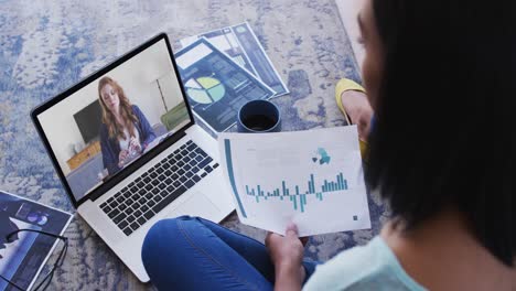 Mixed-race-businesswoman-sitting-on-floor-using-laptop-having-video-call-with-female-colleague