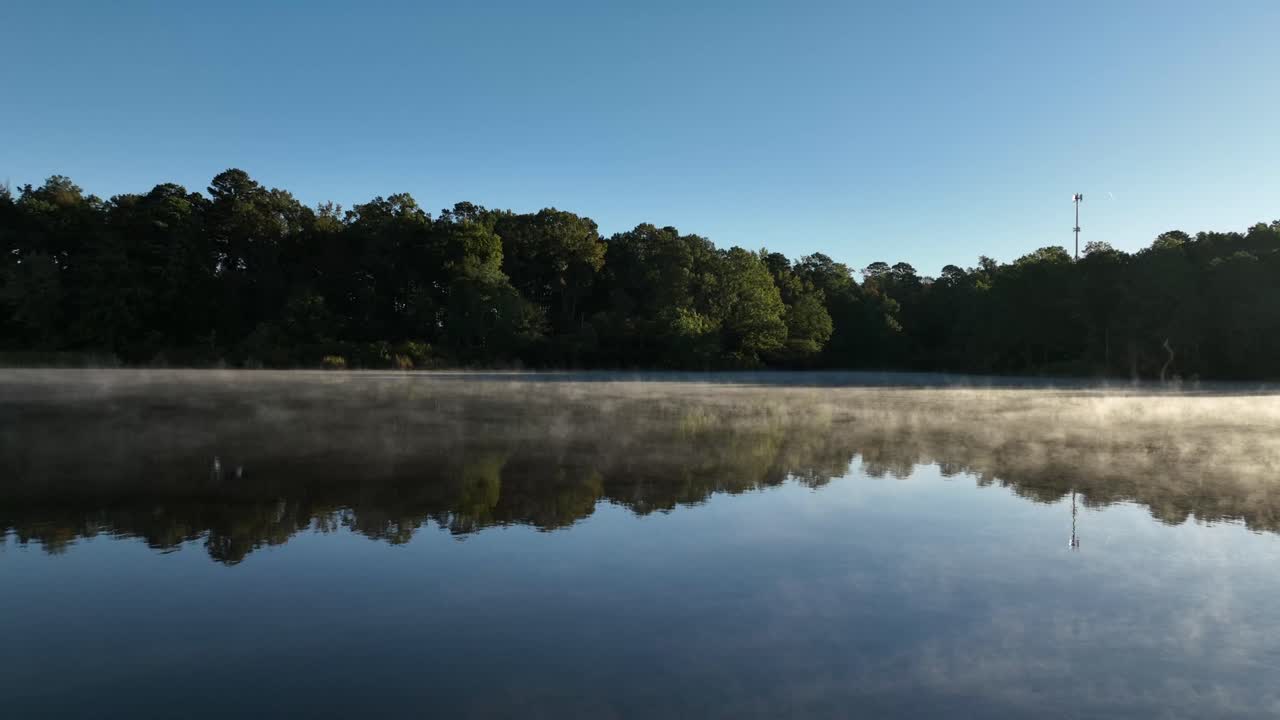 A thin layer of fog drifts across a calm sunrise lake with clear tree reflections