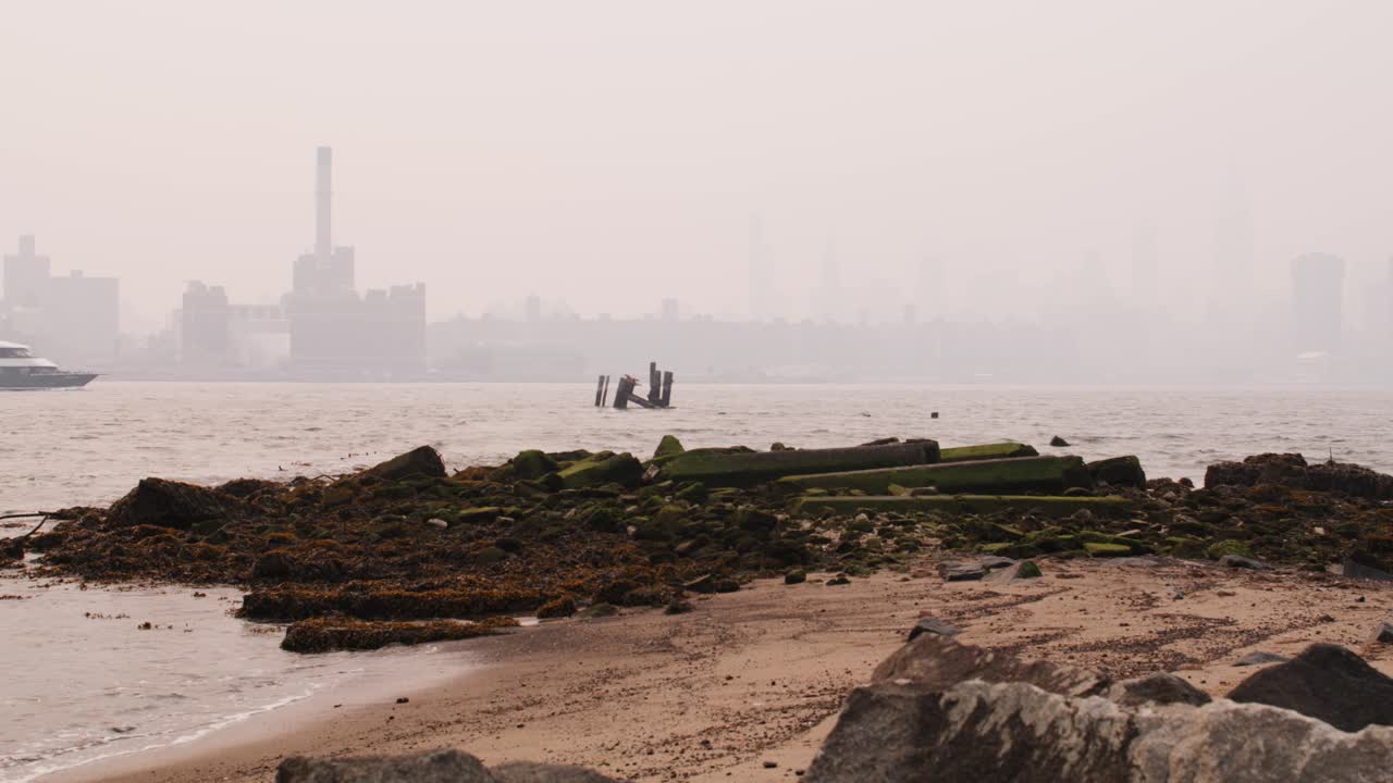 View of Manhattan covered in smoke from wildfires seen from beach on the east river with waves crashing on mossy rocks in the foreground and a ferry crossing frame