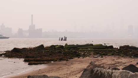 View-of-Manhattan-covered-in-smoke-from-wildfires-seen-from-beach-on-the-east-river-with-waves-crashing-on-mossy-rocks-in-the-foreground-and-a-ferry-crossing-frame