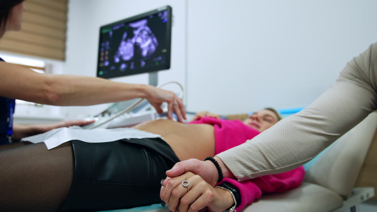 Caucasian man strokes tenderly his wife's hand who is having sonography of pregnancy. Expecting couple at doctor's reception.