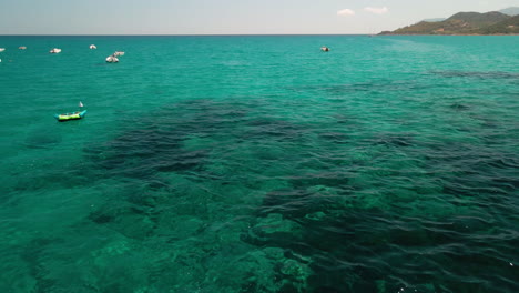 Blue-Green-Waters-Of-The-Mediterranean-Sea-With-Moored-Boats-And-Mountains-In-Background-During-Summer-In-Sardinia,-Italy---aerial-pullback