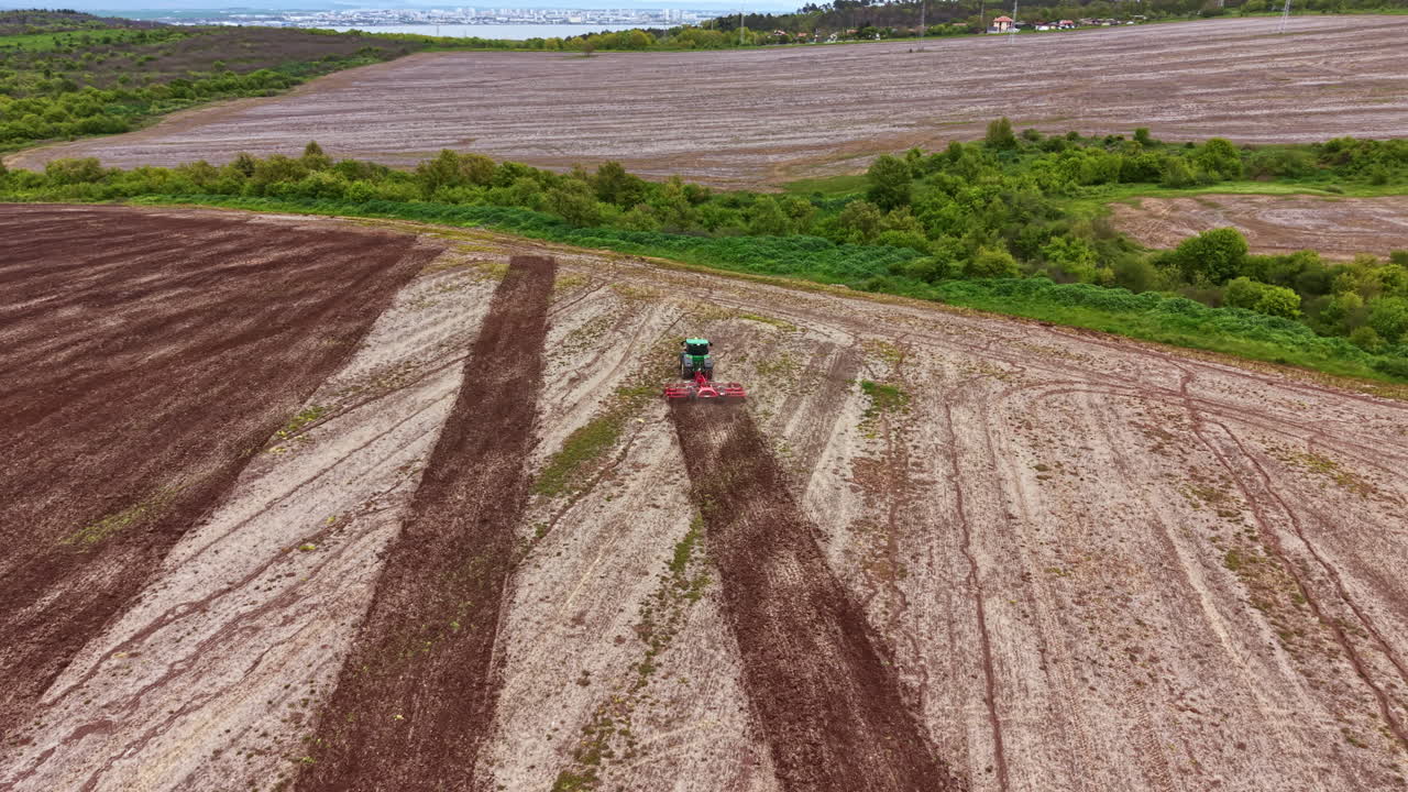 Large tractor works on a field during planting season in a rural area near a city with green hills in the background