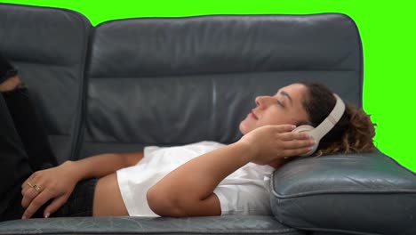 girl laying on couch listening to music with headphones turns to camera, chroma key green screen background