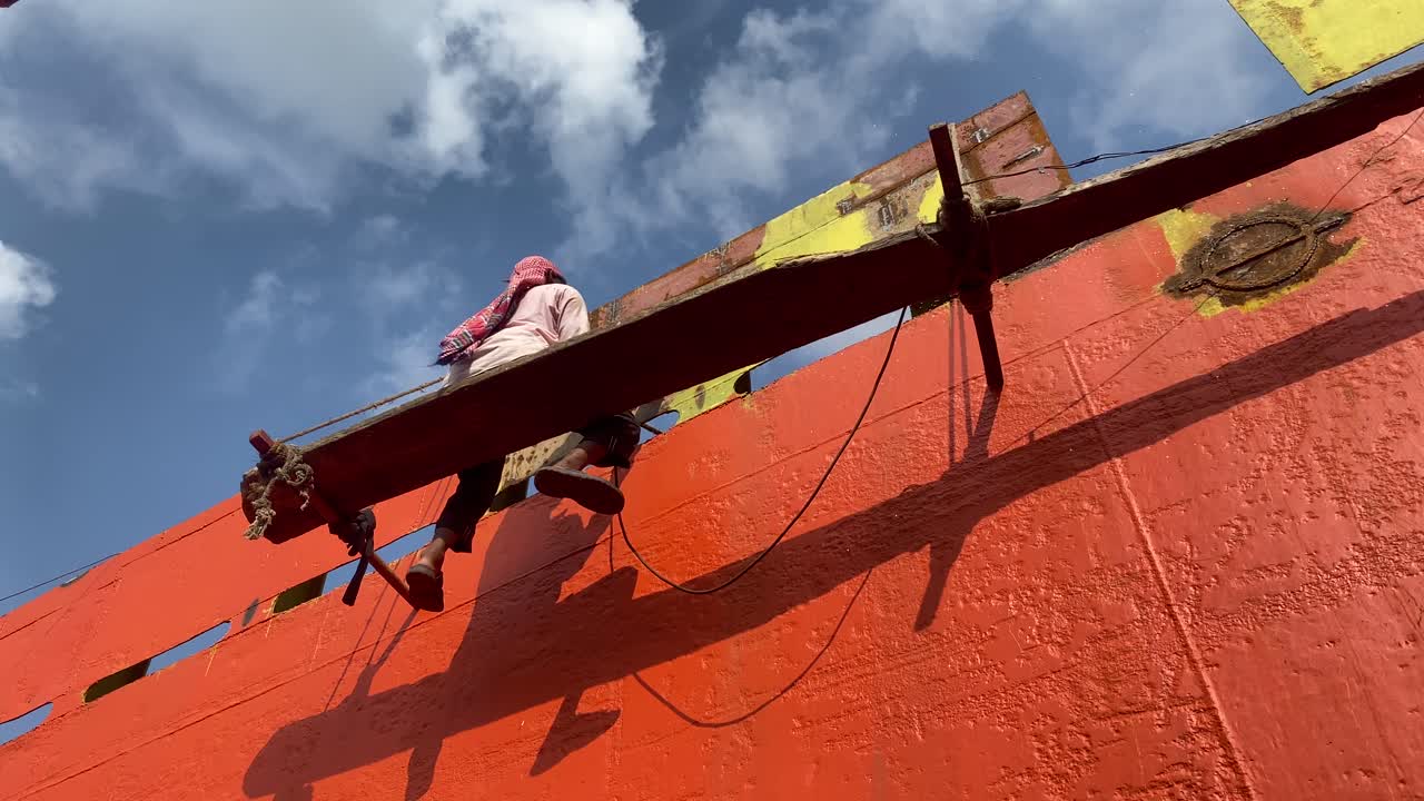 In a third world country, a working person labors on a ship hull, scaling scaffolding to repair and maintain the vessel