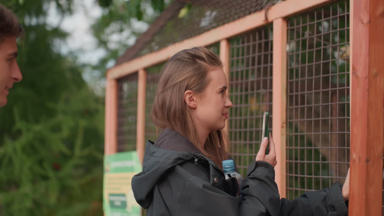 young woman holding phone at enclosure filming animals through wooden mesh fence, casual jacket, candid smile, handheld closeup for social media content, observational park visit with exhibit signage