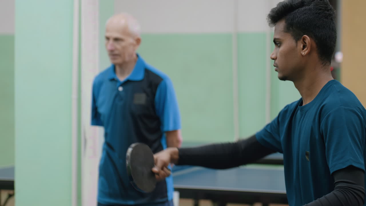 Senior coach in blue shirt attentively observes young player holding paddle at table tennis table, focusing on technique and preparation during indoor sports training session