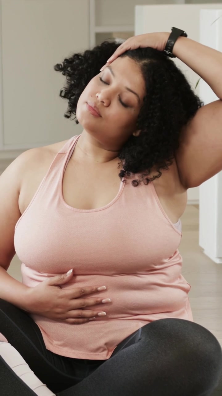 Video verticale: Una donna con canotta rosa mette le mani sulla pancia, guidando il respiro per calmarsi sul tappetino, con un orologio.