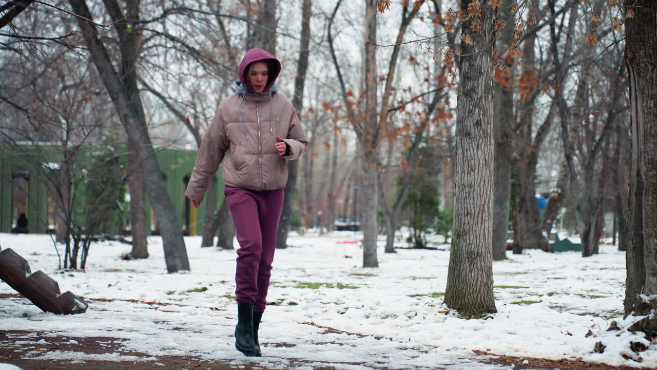 Uma senhora com traje de inverno se aquece movimentando-se em um parque ao ar livre coberto de neve, rodeada por árvores. Vestindo moletom com capuz, botas e luvas, ela pisa com cautela no chão escorregadio enquanto se adapta ao tempo frio.